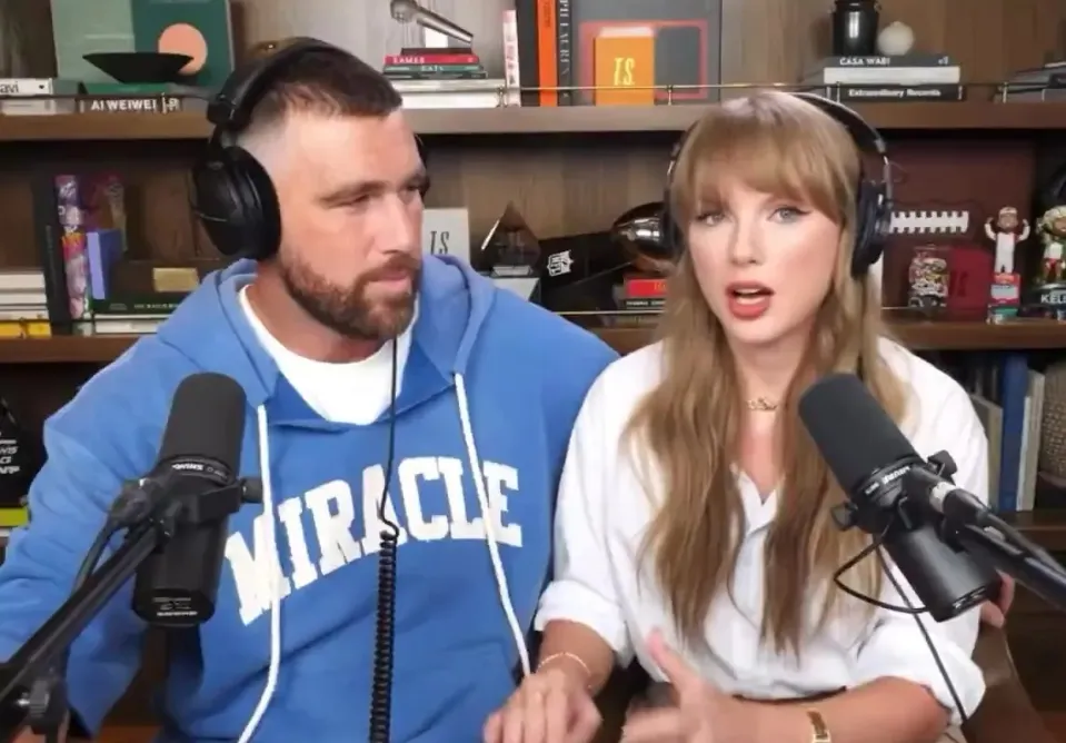 Travis Kelce in a blue sweatshirt and Taylor Swift in a white shirt, both wearing headphones and speaking into microphones on the New Heights podcast.