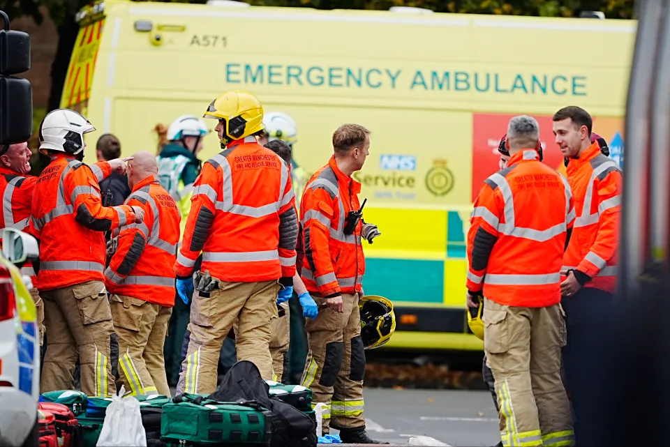 Emergency services personnel, including Greater Manchester Fire and Rescue Service, at an incident scene with an ambulance in the background.