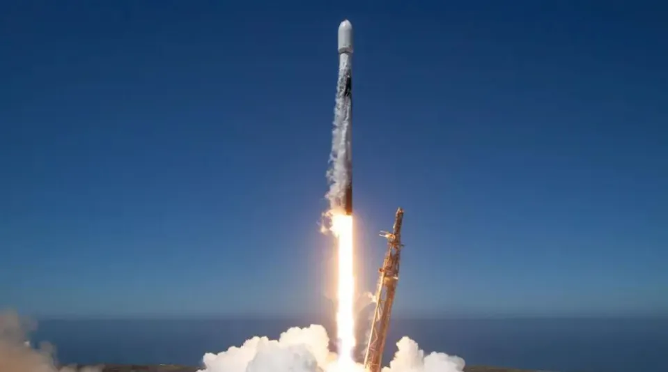 A rocket launching into a clear blue sky, with bright orange flames and white smoke emanating from its base.