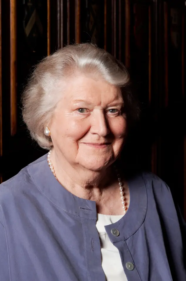 Portrait of Patricia Routledge with short grey hair, wearing a grey shirt and pearl necklace.