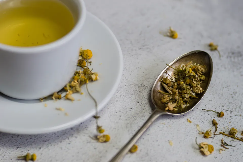 A high-angle view of a white cup of chamomile tea and a spoon with dried chamomile flowers.