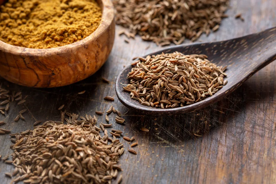 Caraway seeds on a wooden spoon next to a bowl of ground cumin.