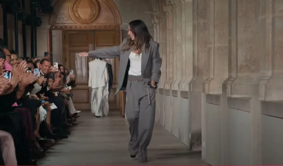 Victoria Beckham walks the runway of her fashion show in a gray suit, waving to the audience.