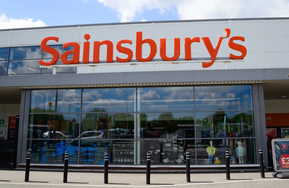 Exterior of a Sainsbury's supermarket with its large orange logo, featuring reflective windows showcasing merchandise and reflections of the sky and surroundings.