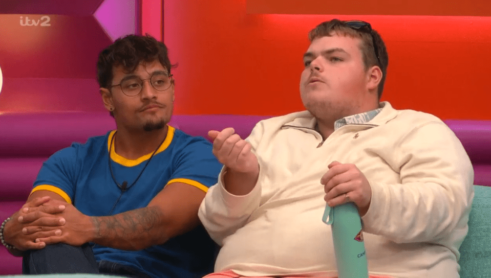 Two men sitting on a couch, one speaking and holding a bottle while the other listens.