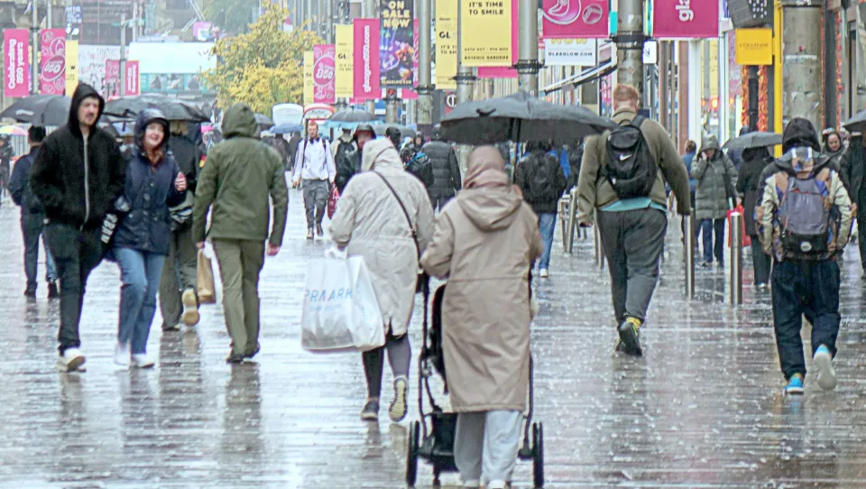 People walking on a wet city street, some holding umbrellas, with banners advertising "Glasgow 850" and sales.