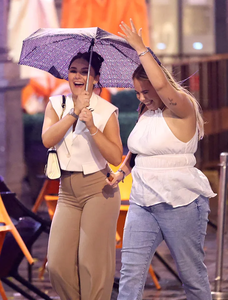 Two young women walking in the rain, one holding an umbrella and the other with her arm raised.