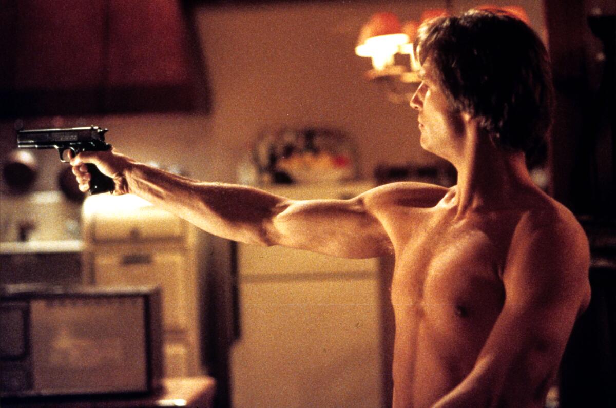 A shirtless man holding up a gun in a kitchen.