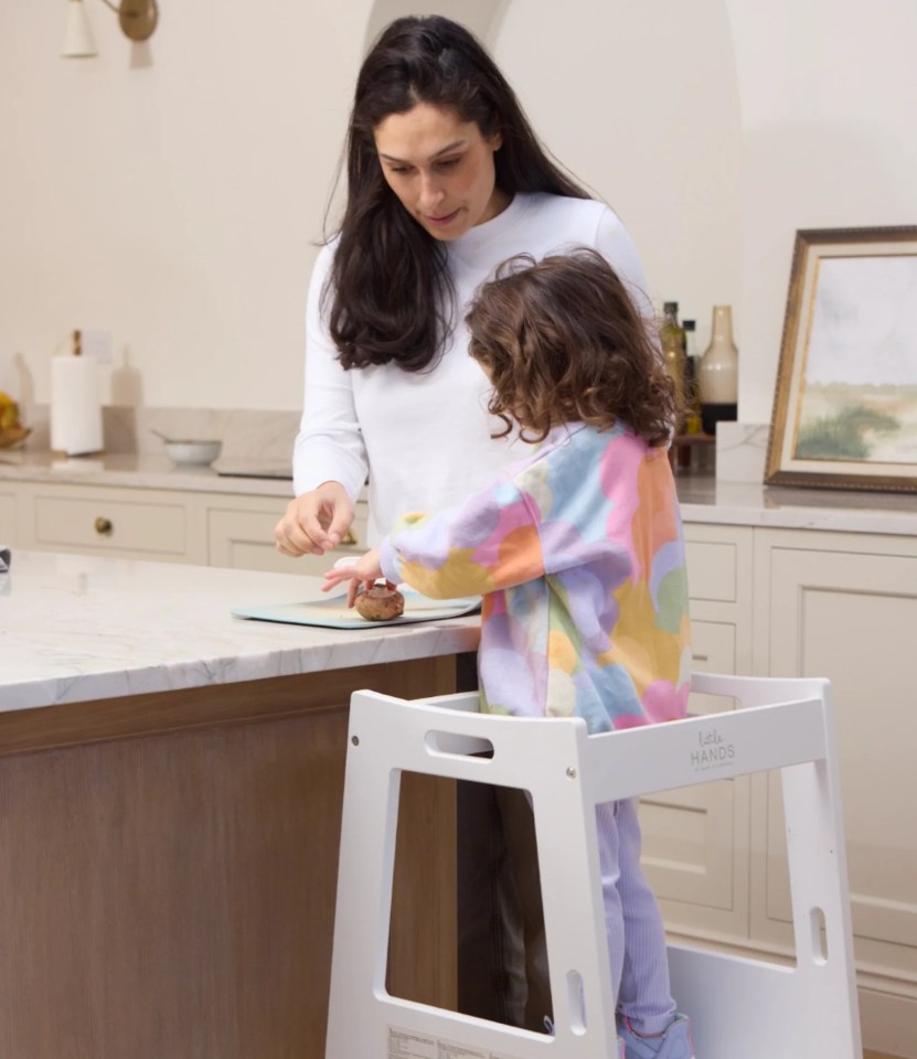 A woman helps a young child in a toddler tower prepare food on a kitchen counter.