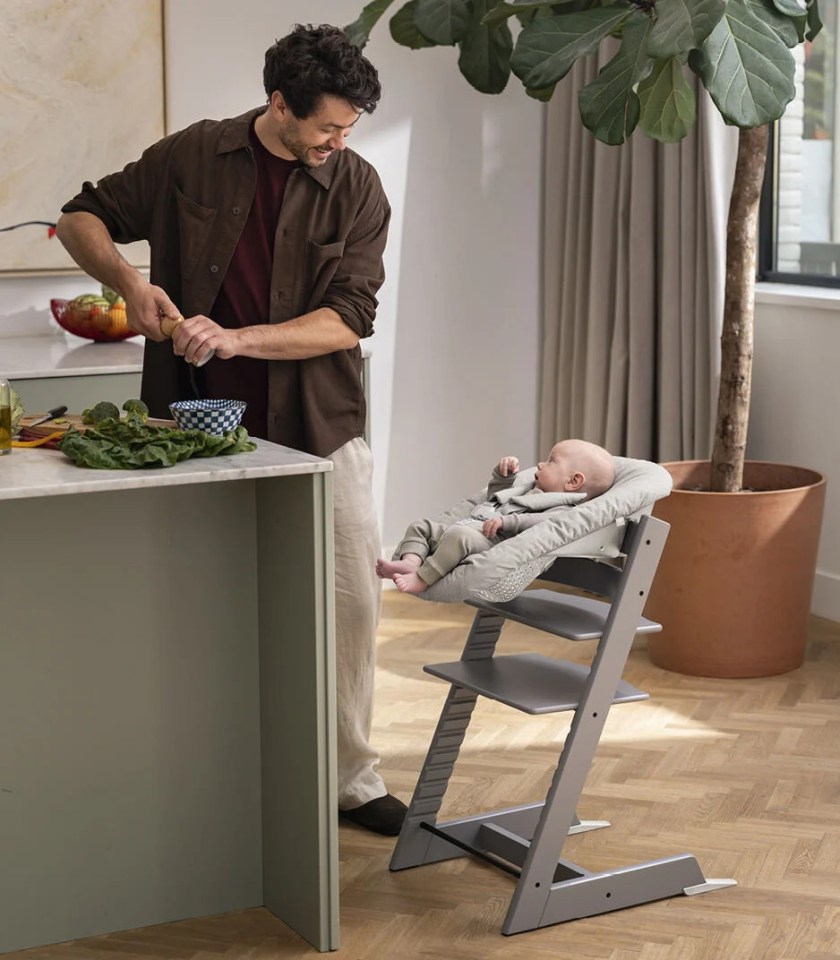 A man grinds spices while a baby sits in a gray newborn set and looks at him.