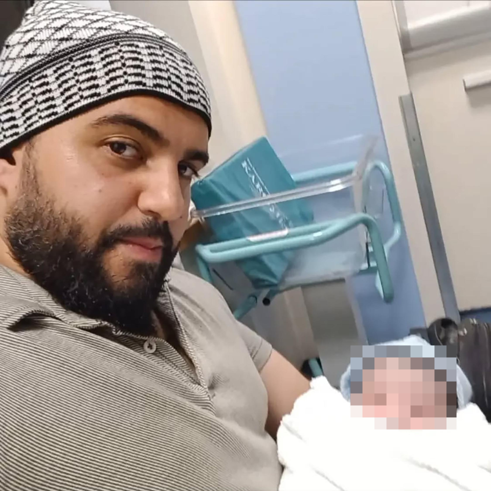 A man in a patterned cap holds a baby whose face is pixelated.