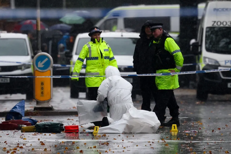 A forensic team member in a white suit works on the ground at a crime scene while two police officers in high-visibility jackets stand nearby, all under falling rain.