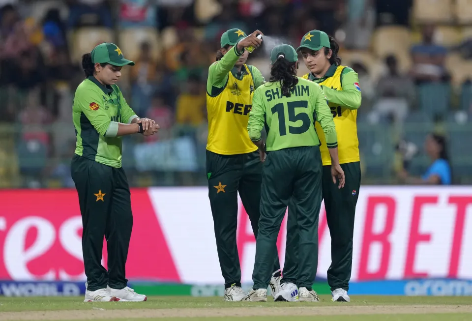A female cricketer sprays another with insect repellent, while two teammates watch, all wearing green and yellow uniforms.