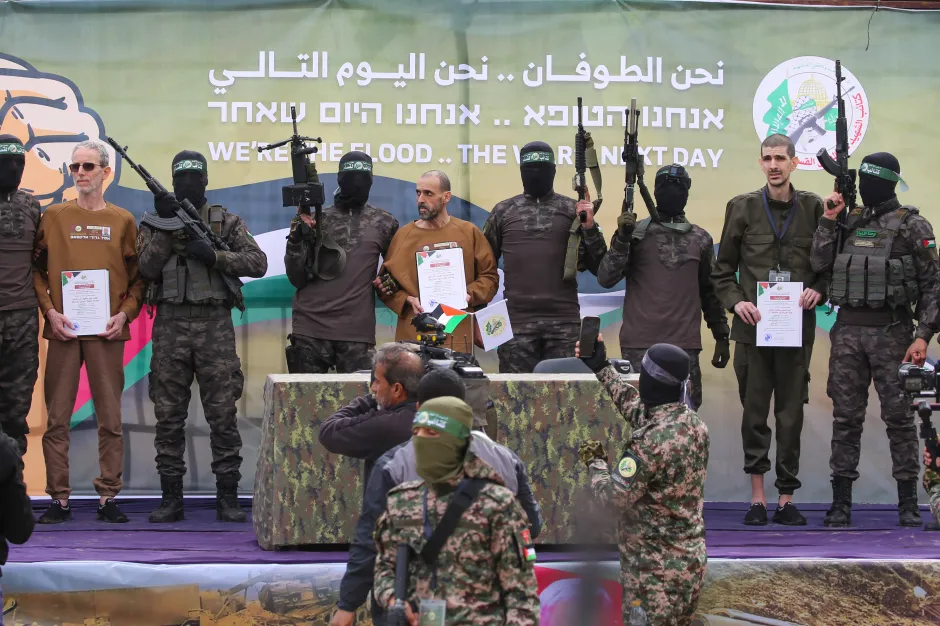 Several armed men in military attire stand with three civilians holding documents, in front of a banner that reads "WE'RE THE FLOOD.. THE NEXT DAY" in English, Arabic, and Hebrew.
