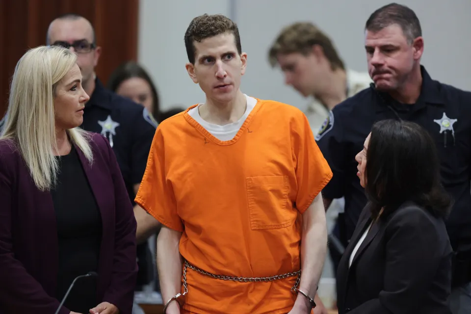 Bryan Kohberger in an orange jumpsuit and handcuffs during a sentencing hearing, flanked by two female lawyers and two male officers.