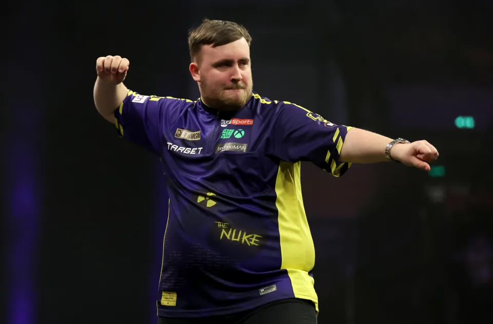 Darts player Luke Littler celebrates, arms raised, wearing a purple and yellow jersey.
