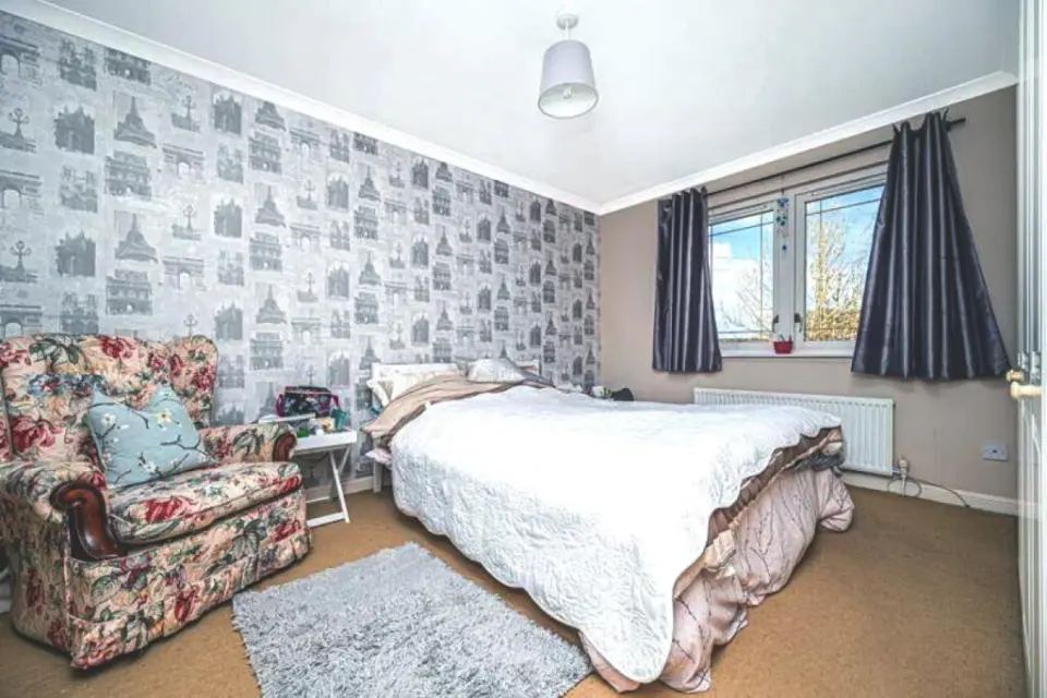 A bedroom with one wall covered in wallpaper depicting historical buildings, a double bed, a floral armchair, and a window with dark curtains.