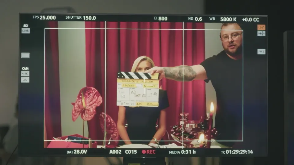 A film set monitor showing a blonde woman holding a clapperboard in front of a red curtain.
