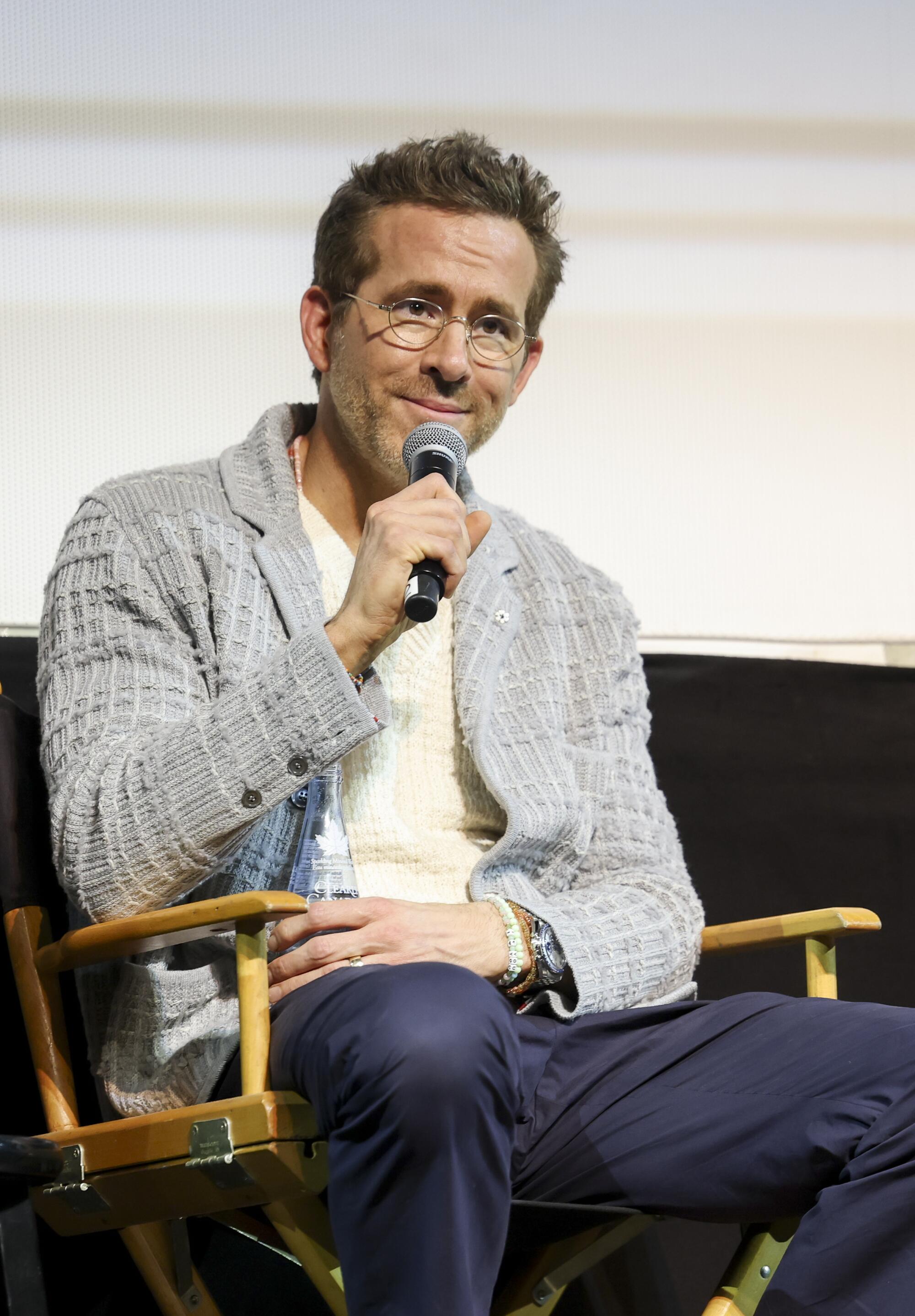 A man in glasses, a gray sweater and jeans sits on a directors chair with a microphone near his mouth.