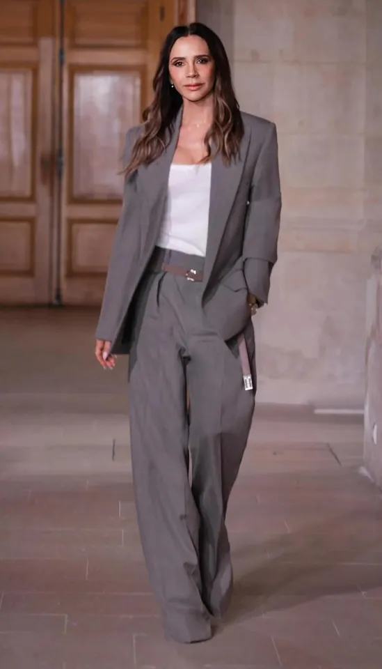 Victoria Beckham on a runway wearing a gray pantsuit with a white top and a brown belt.