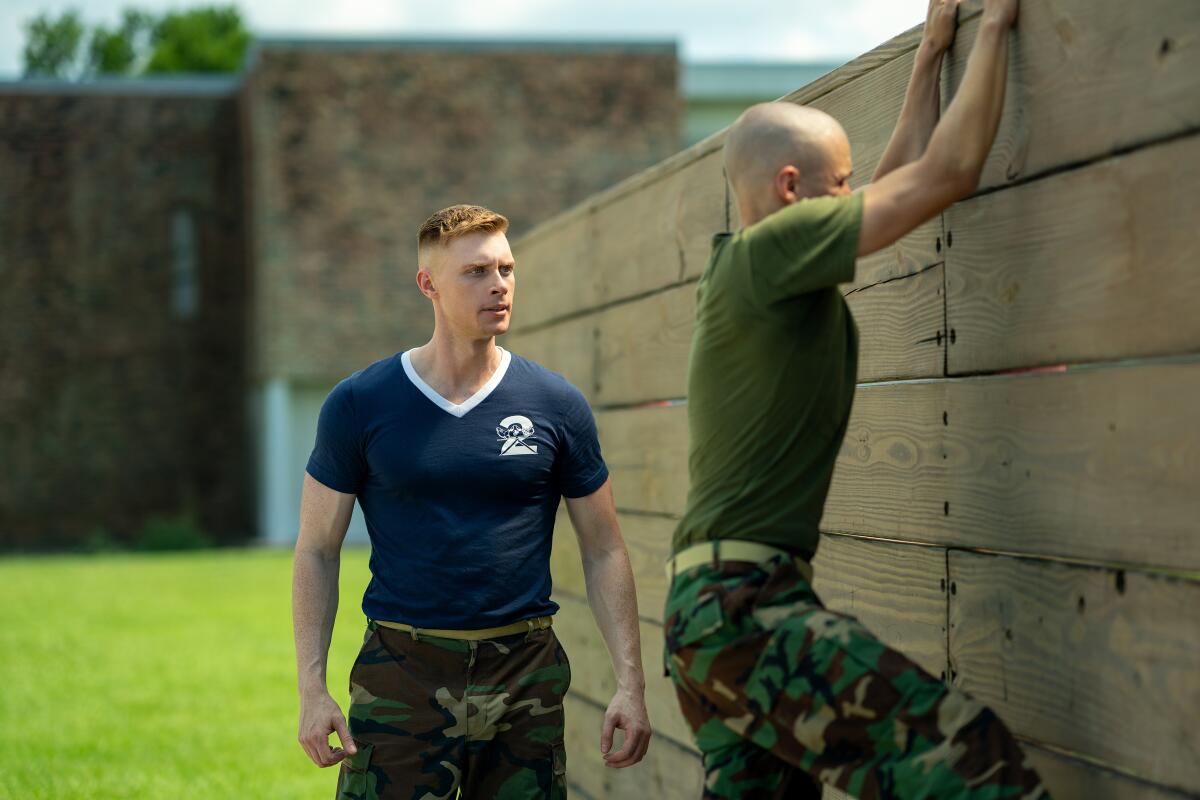 A man in a blue T-shirt and camouflage pants watches a man try to scale a wooden fence.