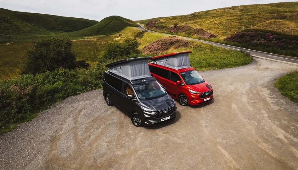 Two Landseer camper vans, one red and one black, with pop-top roofs on a gravel path overlooking a road and hills.