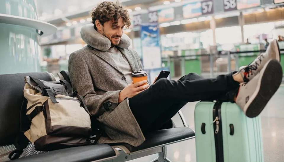 A man in an airport waiting area, wearing a neck pillow and coat, uses his phone while holding a coffee cup, with his feet resting on a light blue suitcase.