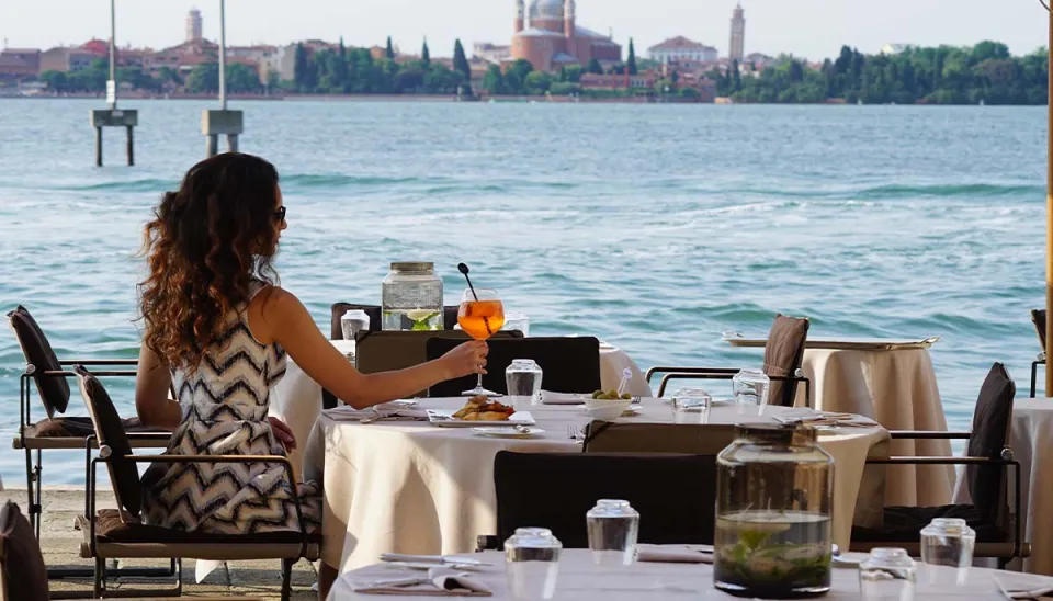 A woman sitting at a table with an orange cocktail in hand, overlooking the sea with a city in the background.