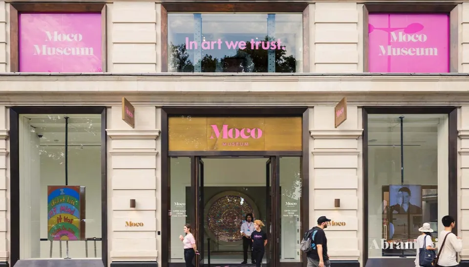 Exterior of the Moco Museum with "In art we trust" written on a window and pink Moco Museum signs.