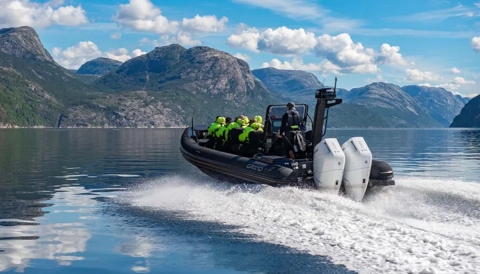 A boat with passengers in yellow life vests speeds across a fjord with mountains in the background.