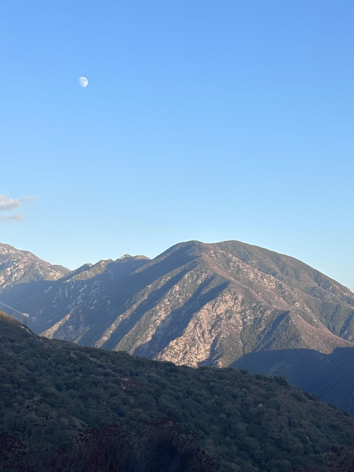 The tiny white moon in the bright blue sky over mountains speckled in red, green and brown plants.