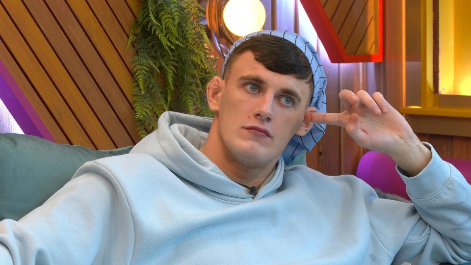 Cameron B, a white man in a light blue hoodie and light blue head covering, sits on a couch with his finger in his ear.
