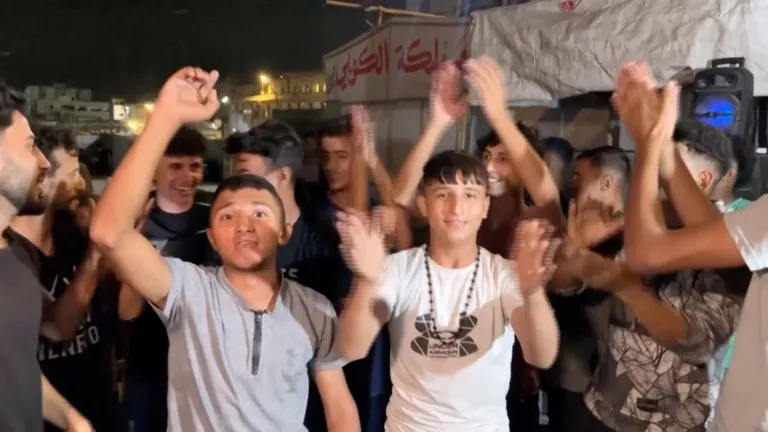 A group of men and boys celebrating and clapping, with Arabic writing on a sign in the background.