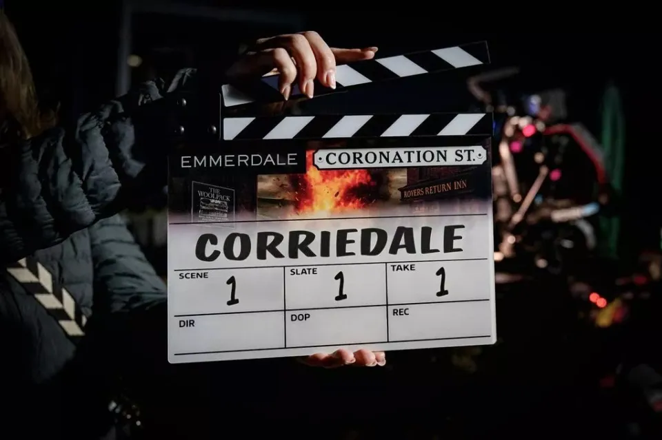 A person holding a clapperboard for a joint "Corriedale" episode of Emmerdale and Coronation Street, showing "Scene 1, Slate 1, Take 1".