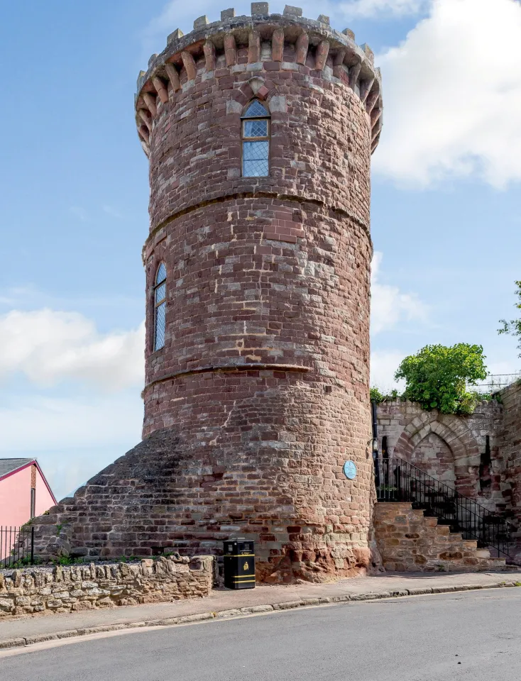 The Gazebo Tower, a small, round, red brick castle with crenelated battlements, a single arched window, and a blue plaque on its side, next to a road.
