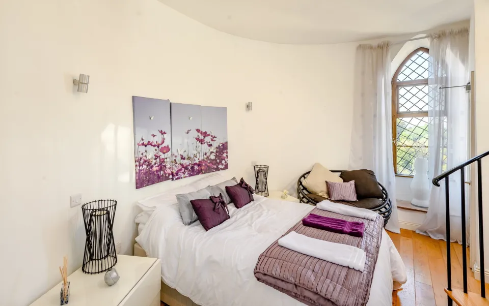 A bedroom with white walls, a bed with purple pillows and blankets, a dark brown chair, and a leaded glass window with a view of greenery.