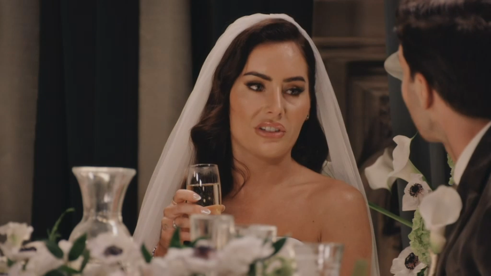 Leisha, wearing a wedding veil and holding a glass of champagne, looks at Reiss.