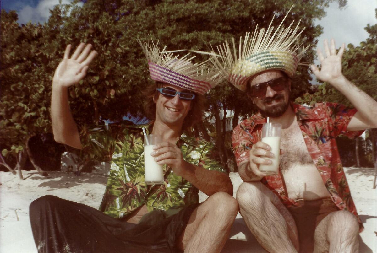Two men in basket hats and shades smile at the camera with tropical drinks.