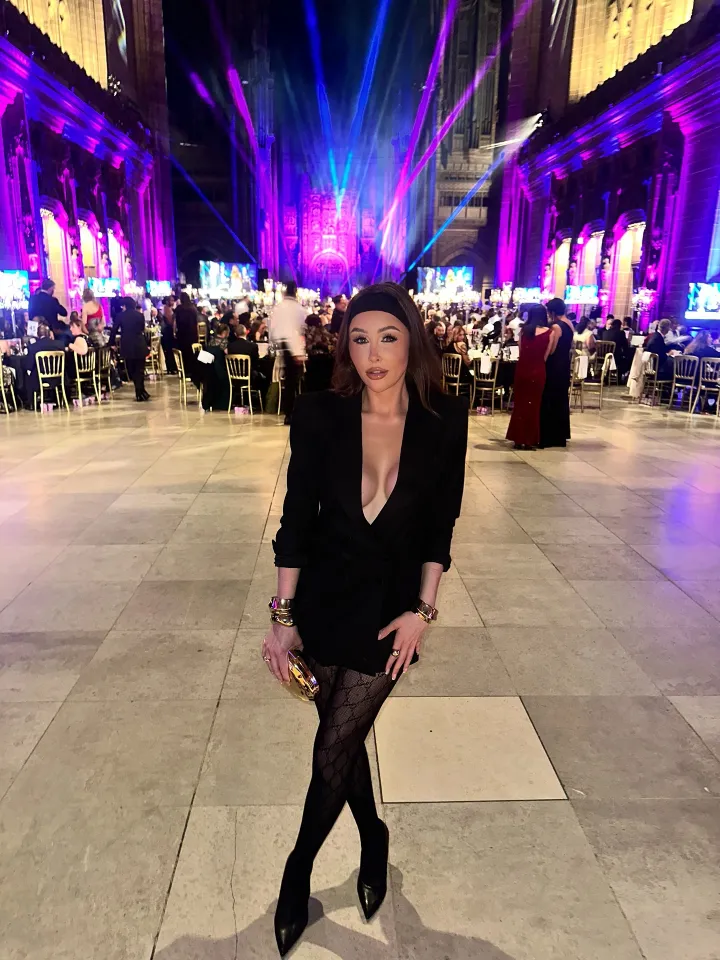 Ella Morgan poses in a black blazer, patterned tights, and heels at an event with purple and blue lighting.