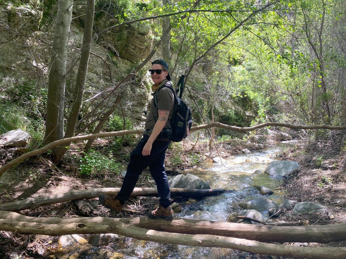 A smiling hiker balances on multiple logs stretched over a narrow creek.
