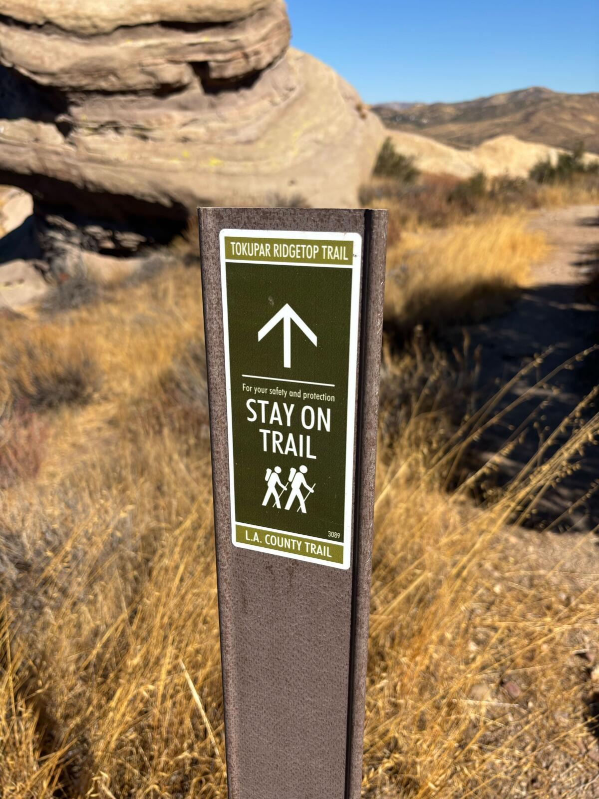 A narrow trail sign directing hikers to stay on trail in a desert landscape.