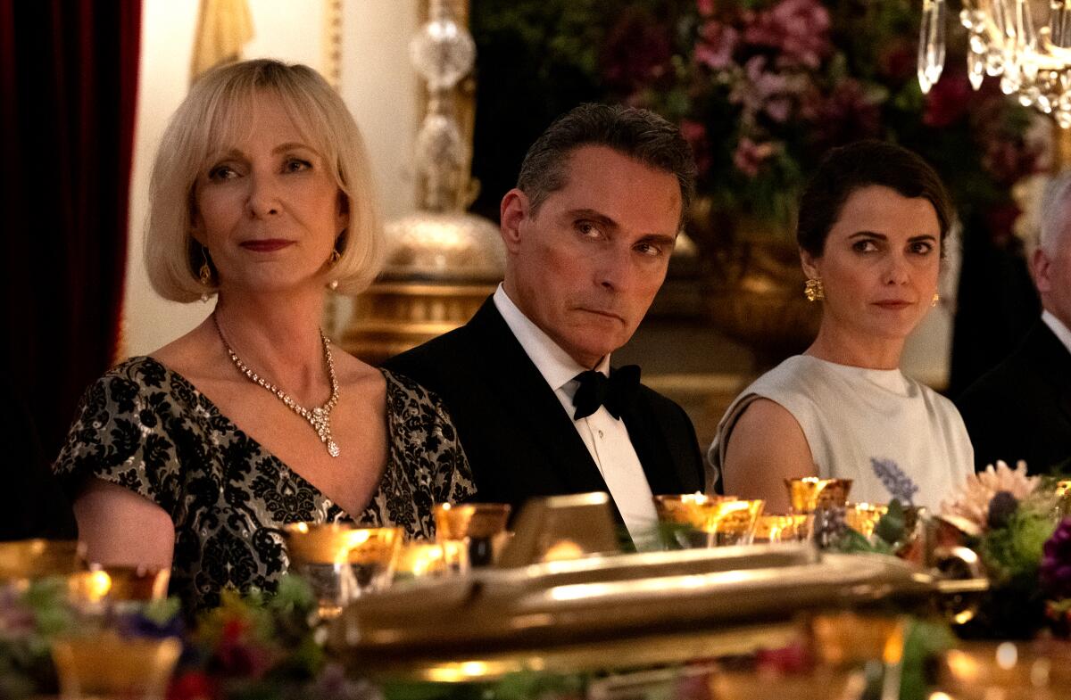 Three well-dressed people -- a man sitting between two women -- gaze to their right while seated for dinner