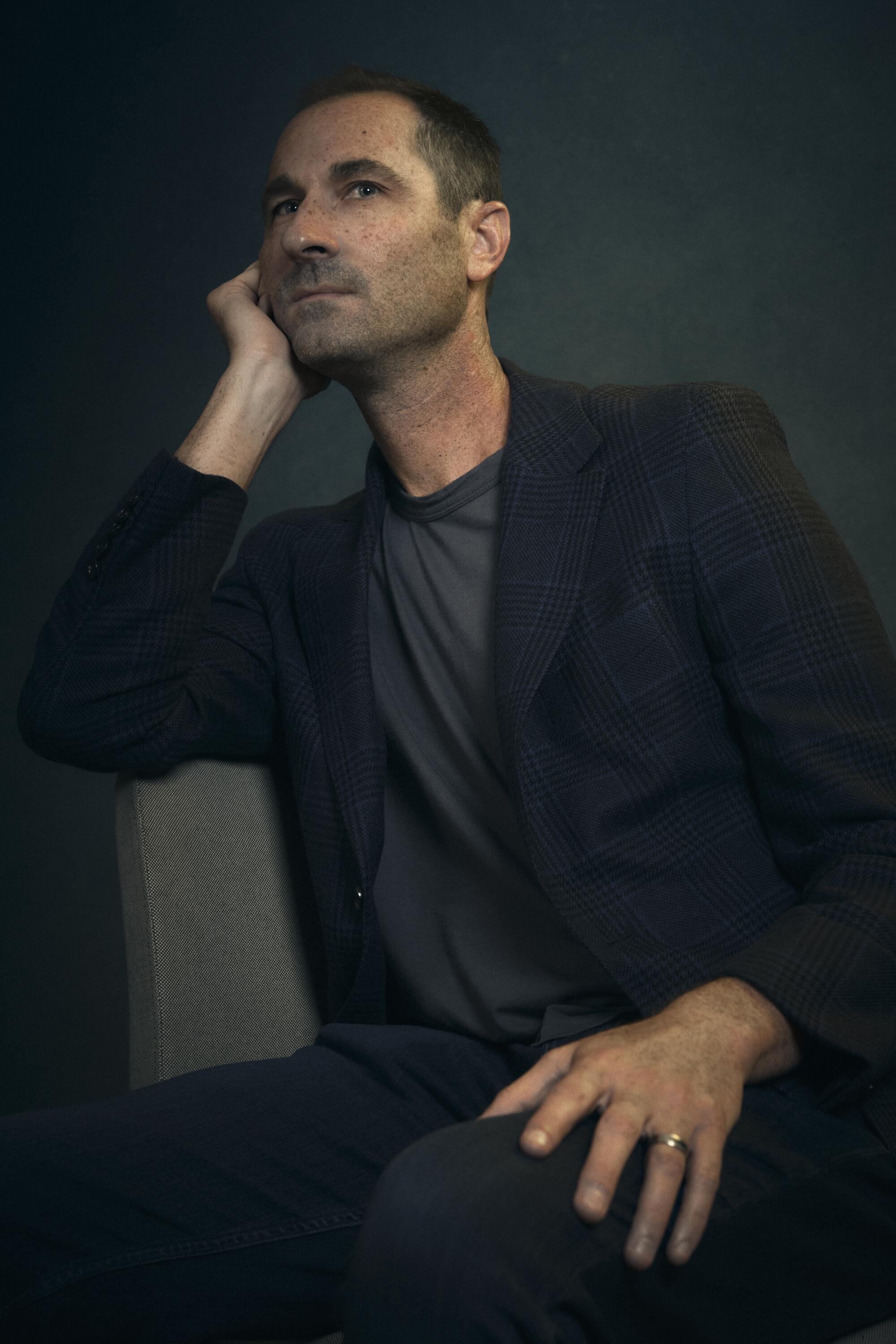 A man in a suit jacket poses for a photo in a chair.