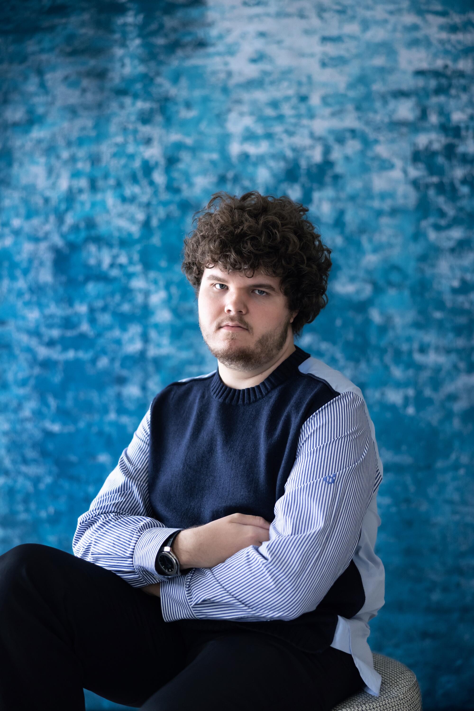 A young man sits in front of a blue backdrop with his arms crossed.