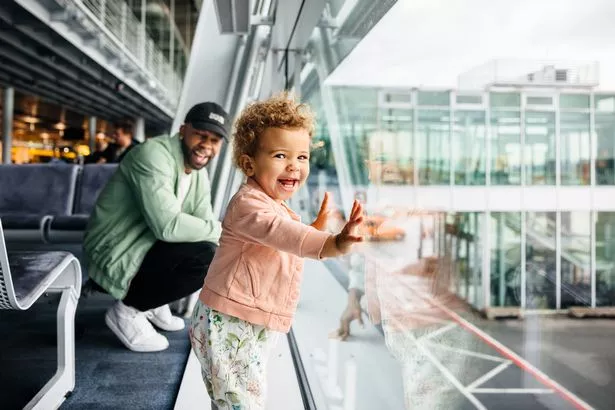 A joyful toddler girl presses her hands against the airport glass as her laughing father crouches behind her, both enjoying their playful time together.