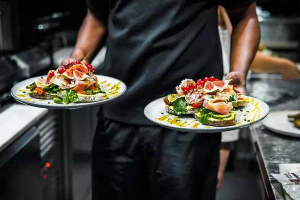In a bustling kitchen, a chef presents two plates of beautifully garnished avocado toasts, ready to be served to awaiting customers.