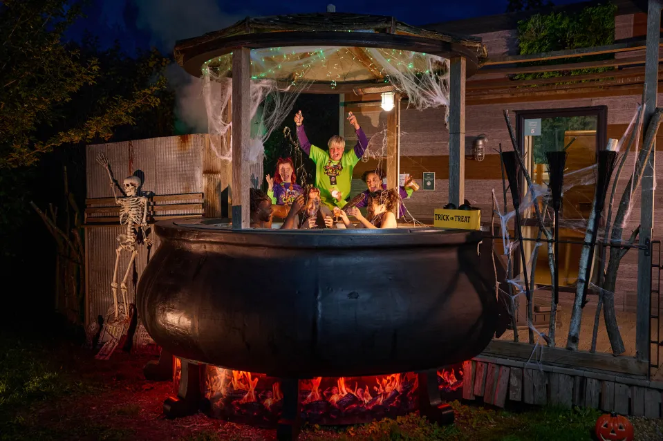 Five people enjoying a Halloween-themed hot tub, designed to look like a witch's cauldron, at night, with a skeleton and cobwebs decorating the scene.