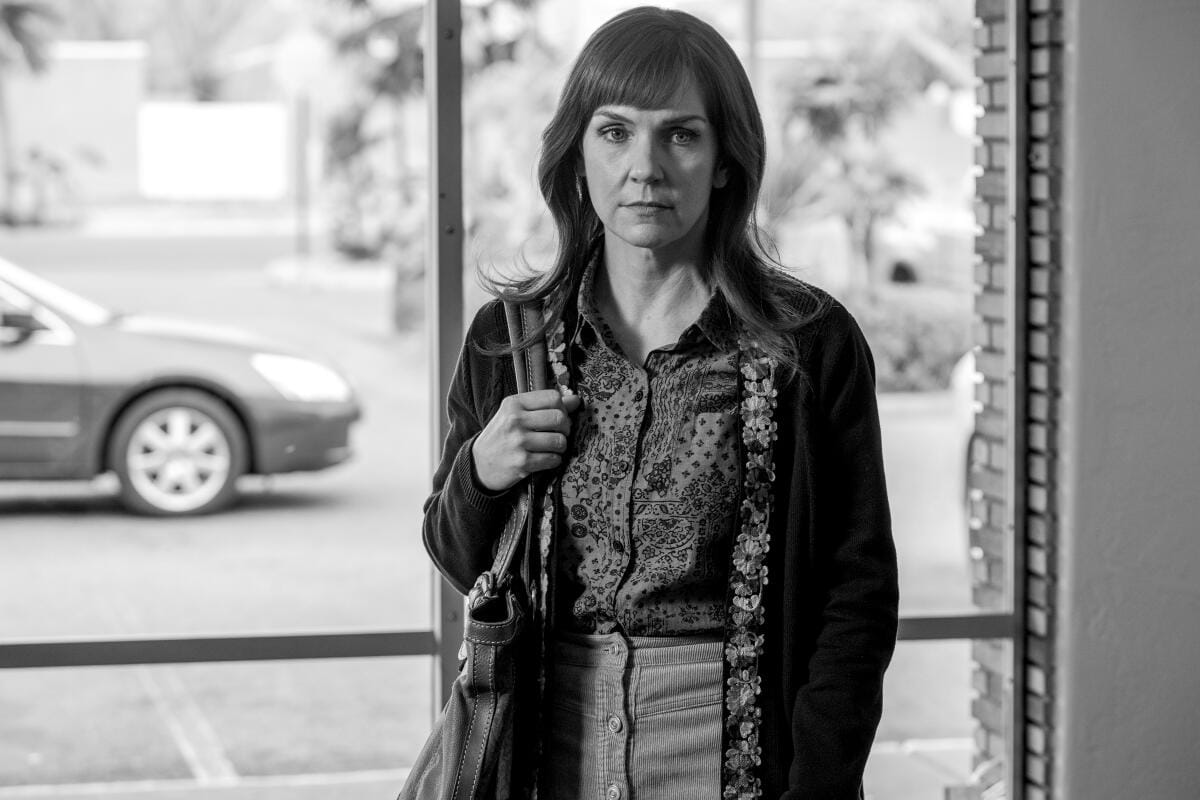 A woman with shoulder-length hair and bangs holding a bag on her shoulder stands near a window.