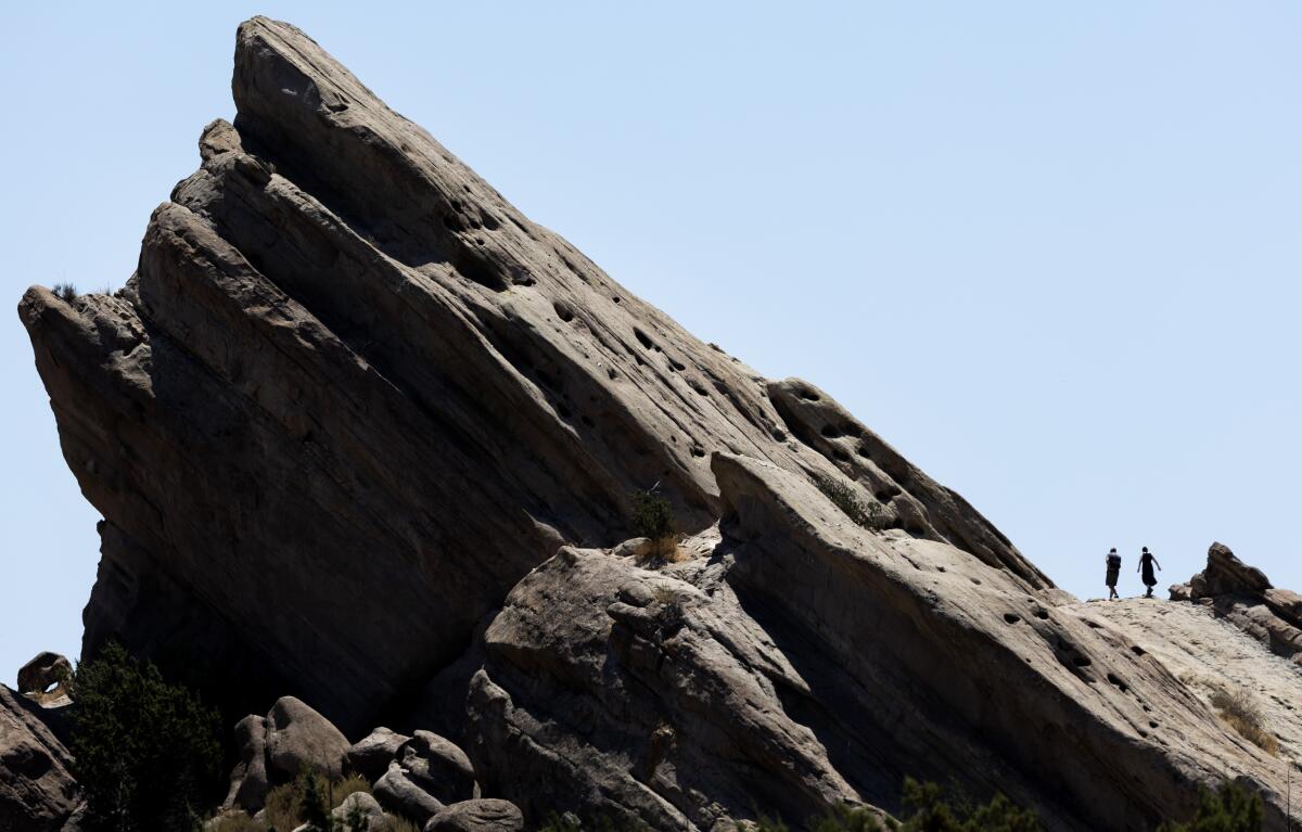 Two people explore near massive slabs of rock that sit at an angle.