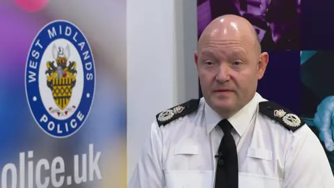 Craig Guildford in a picture. He is in a West Midlands Police office. He is wearing full uniform. He has a white shirt and black tie.